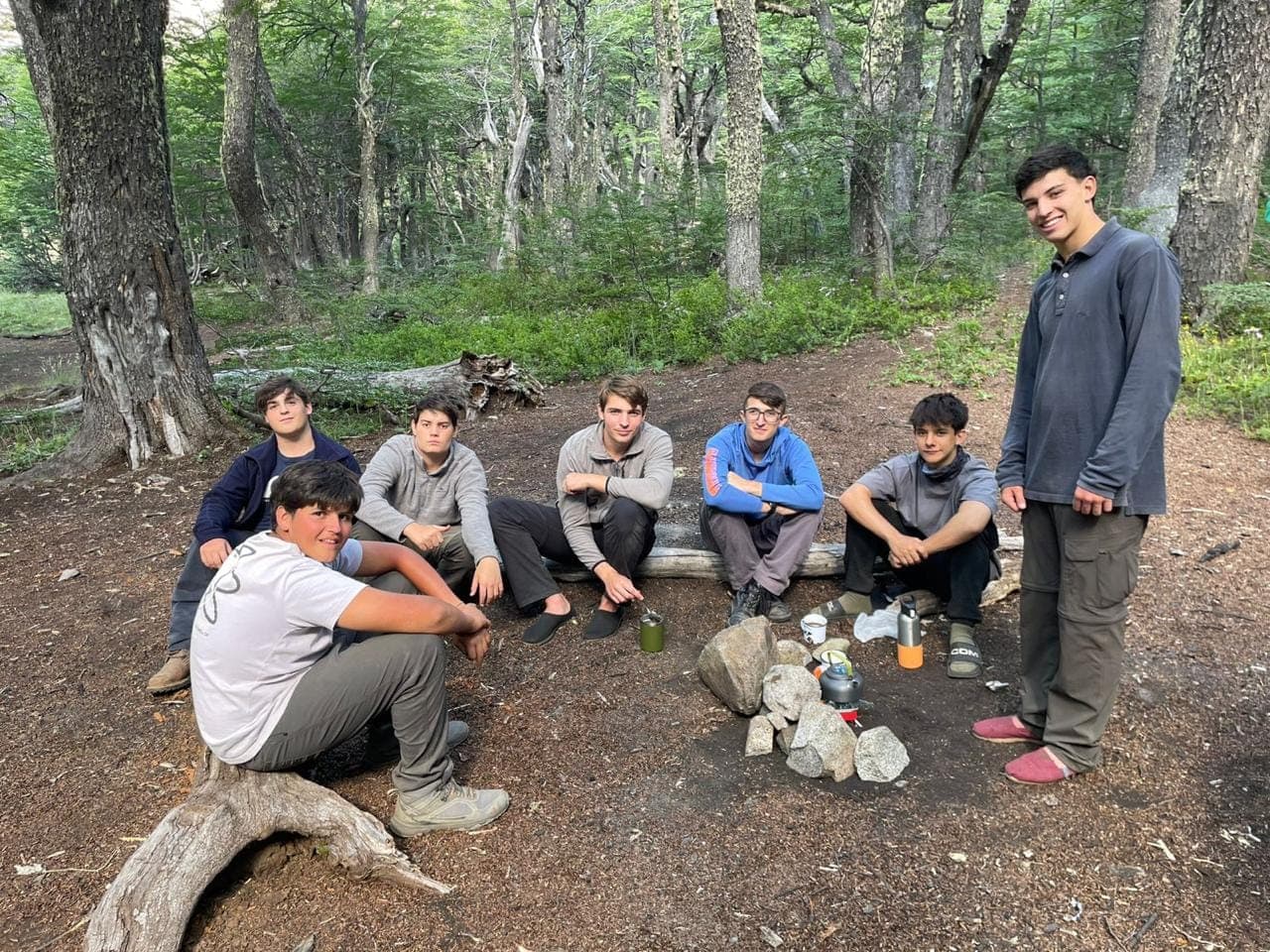 Chicos sentados sobre un tronco en el bosque, con el calentador y los jarros